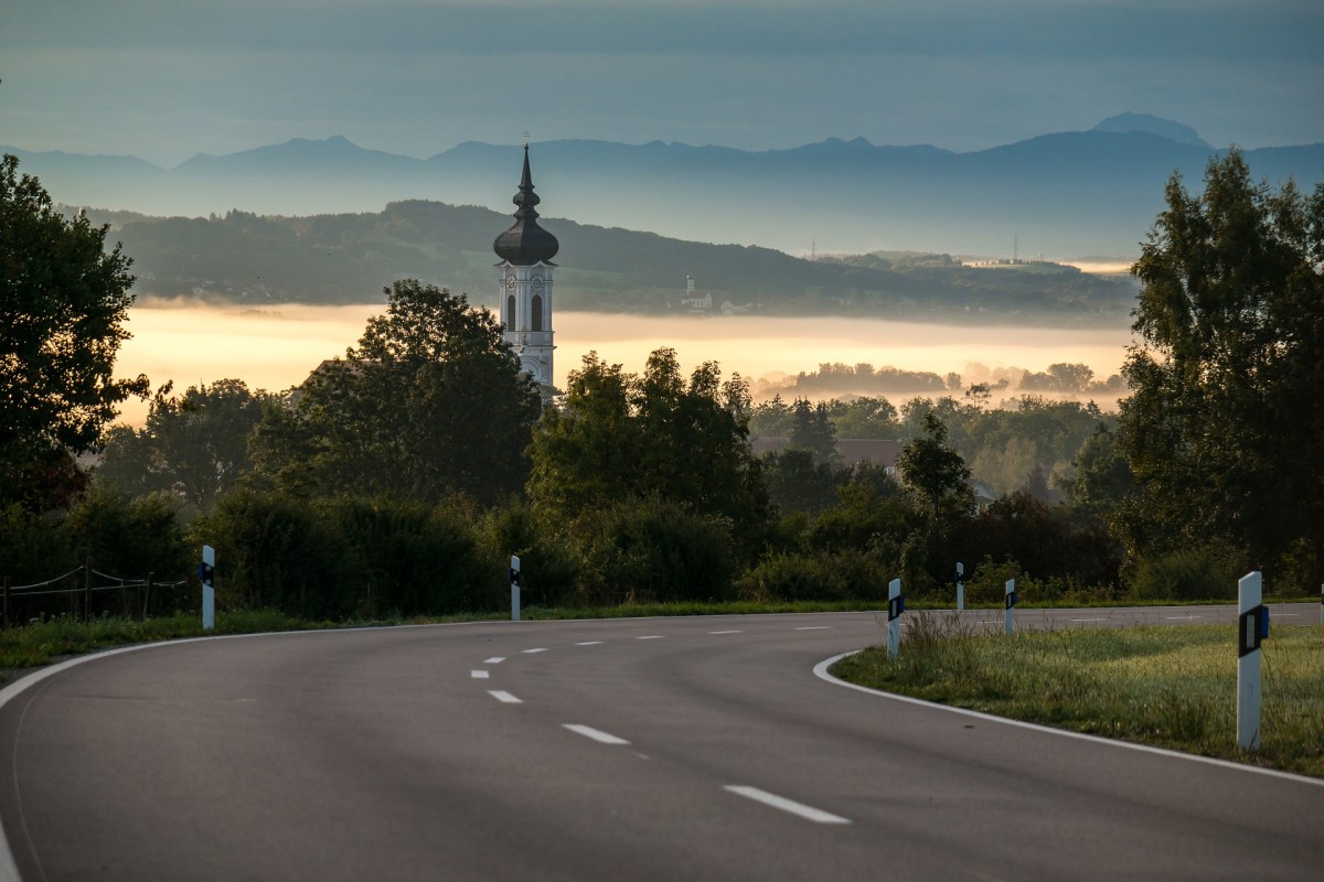 Straße in Bayern mit Kirchturm und den Alpen im Hintergrund